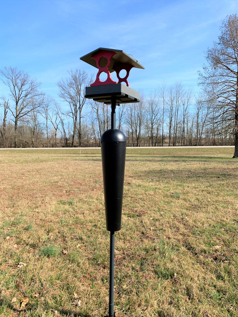 Photograph of a dark gray and red bird feeder in a rural field with trees and a road in the background featuring a unique peaked roof and a cylindrical squirrel baffle with a dark gray post