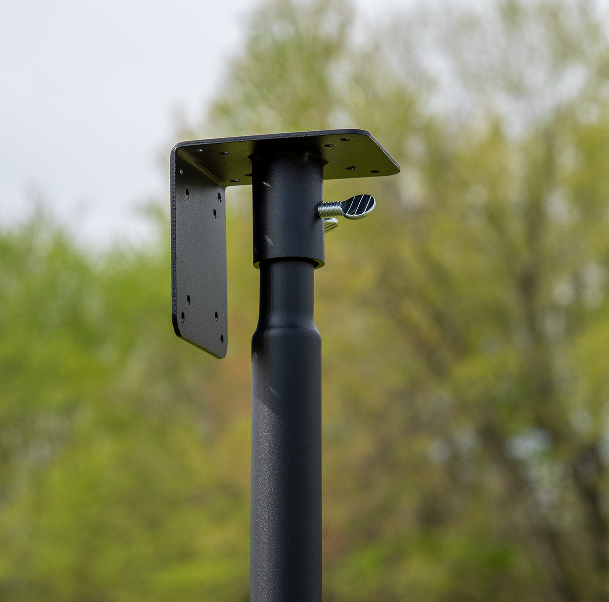 Photograph of a black metal post with a square mounting plate and a silver thumb screw against a blurred background of green trees