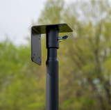 Photograph of a black metal post with a square mounting plate and a silver thumb screw against a blurred background of green trees