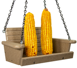 Plastic bird feeder photography showing two bright yellow ears of corn hanging from chains on a miniature taupe-colored swing bench with visible metal hardware and tiny screw holes
