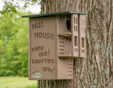 JCS Wildlife brown composite squirrel house affixed to a tree trunk featuring a green roof, carved lettering, and multiple entry holes against a blurred green background
