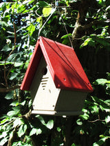 Recycled plastic birdhouse hanging in a lush green garden featuring a vibrant red roof and beige body showcasing ventilation slots and visible screws
