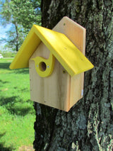 Handmade wooden birdhouse with bright yellow roof and bird-shaped entry hole mounted on a tree trunk in a grassy area near trees
