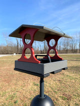 Photograph of a gray and red bird feeder with a unique curved design in a rural setting featuring bare trees and a clear blue sky
