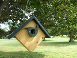 Handmade wooden birdhouse with a dark green roof and bird-shaped entrance hanging from a tree in a grassy park-like setting featuring lush green trees and a partially visible white fence