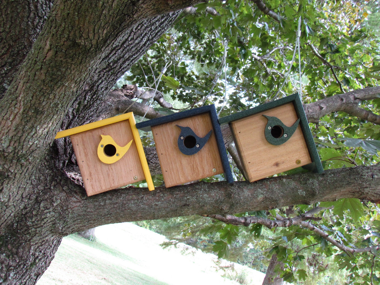 Three handcrafted wooden birdhouses in yellow, dark blue, and dark green hang from a large tree branch with a grassy background showing hints of other trees and featuring bird-shaped entry holes
