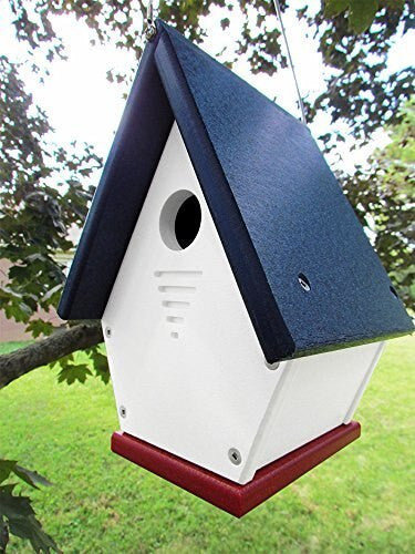 A handcrafted birdhouse with a navy blue roof, white body, and crimson base hangs from a tree in a lush green garden