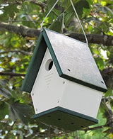 Recycled plastic birdhouse hanging from a tree branch featuring a white body green roof and base drainage holes and a small round entrance