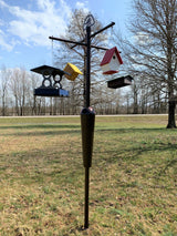 Photograph of a backyard bird feeder station featuring a dark metal pole with hanging birdhouses in red white and yellow a blue and gray feeder and a dark brown seed container against a backdrop of a grassy yard and bare trees