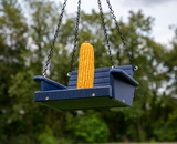 Photograph of a blue plastic bird feeder shaped like a swing hanging outdoors with a bright yellow ear of corn a lush green background and dark metal chains