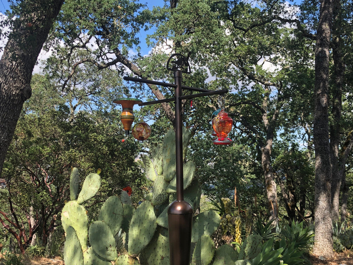Photograph of a multi-tiered hummingbird feeder with colorful glass and ceramic accents amongst prickly pear cacti and tall oak trees in a garden setting
