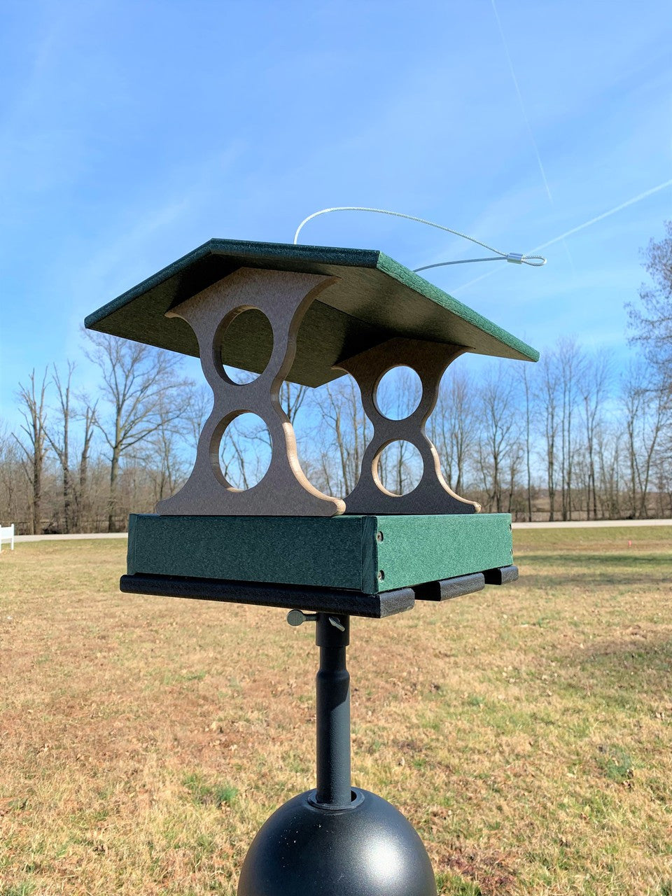 Recycled plastic bird feeder in a grassy field with trees and a road in the background featuring a dark green tray taupe supports and a wire cable
