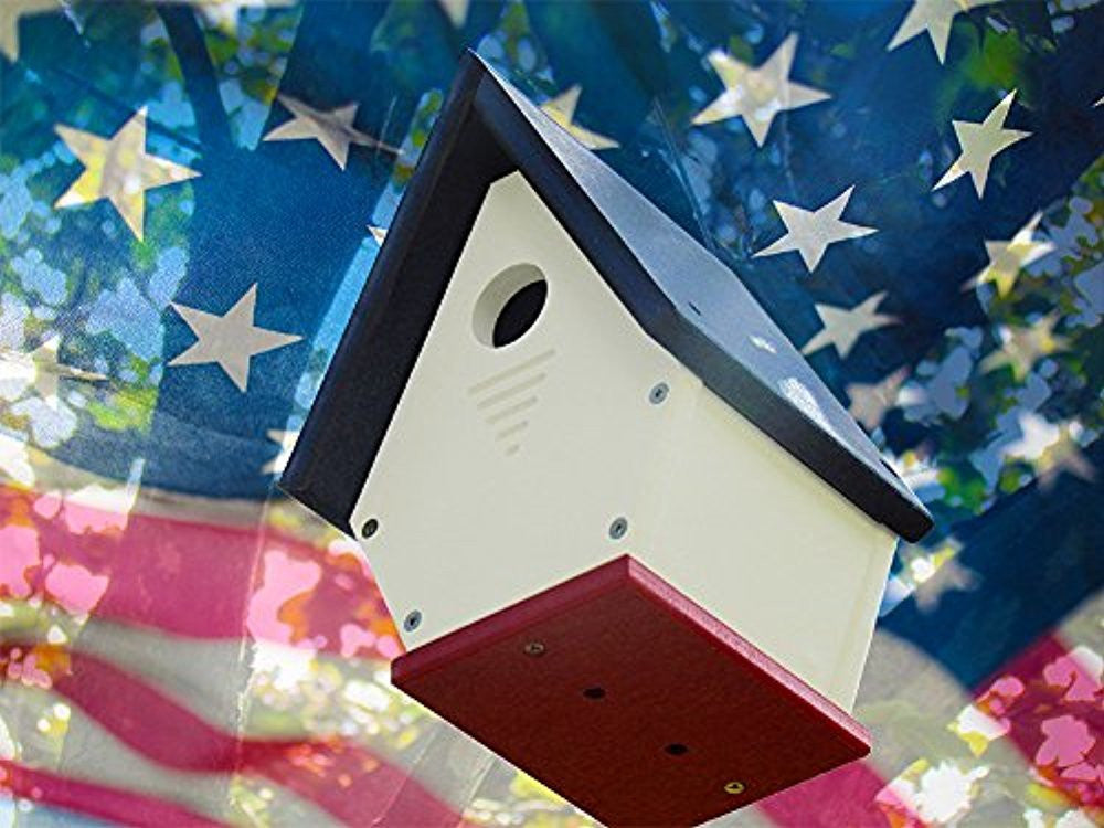 Photograph of a handcrafted birdhouse against a blurred American flag featuring a dark blue roof, white body, and crimson base with visible screws and a circular entry hole