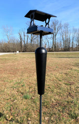 Photograph of a dark blue and black bird feeder with a squirrel baffle set in a grassy field near bare winter trees and a white sign.