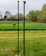 Photograph showing a seven-foot black metal post in a grassy field with a vibrant green wheat field and distant trees in the background, featuring a small square security camera mount, a few yellow wildflowers, and a contrasting patch of lighter green grass.
