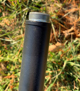 Close-up photo of a black metal pole with a silver threaded top against a blurred background of green and brown grass showing textured paint and subtle light reflections
