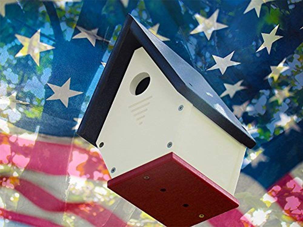 Photograph of a handcrafted birdhouse with a dark blue roof, off-white body, and crimson base against a blurred American flag featuring prominent stars and stripes
