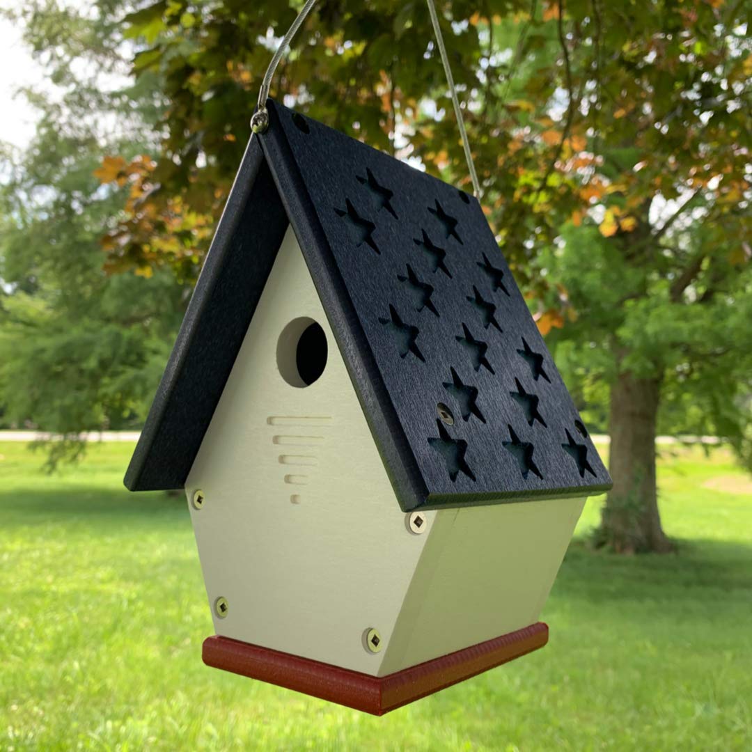 Handcrafted wooden birdhouse with a navy blue star-studded roof, off-white body, and red base hanging outdoors against a lush green lawn and trees
