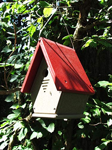 Photograph of a red and beige birdhouse hanging from a tree in a garden with vibrant green leaves and a visible tree trunk