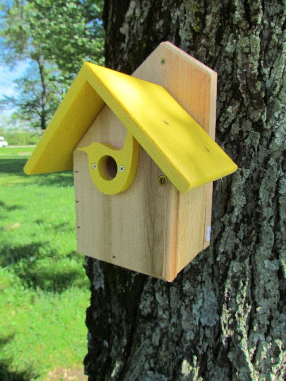 Handcrafted wooden birdhouse with a bright yellow roof and bird-shaped entry hole mounted on a lichen-covered tree trunk in a lush green field

