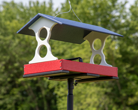 Recycled plastic bird feeder with red tray, white supports, and a navy blue roof outdoors amidst green trees