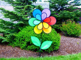 Photograph of a colorful, rainbow wind spinner flower in a garden with evergreen trees and vibrant green shrubs exhibiting bright red, yellow and blue petals