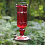 Red antique glass hummingbird feeder with brushed copper base and a hummingbird feeding from one of the decorative ports
