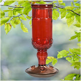 Red antique bottle hummingbird feeder with decorative copper base and metal feeding ports in a lush outdoor setting