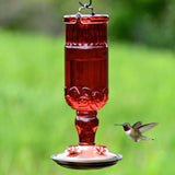 Red antique glass hummingbird feeder with copper base and four feeding ports hanging outdoors with a hummingbird drinking nectar.