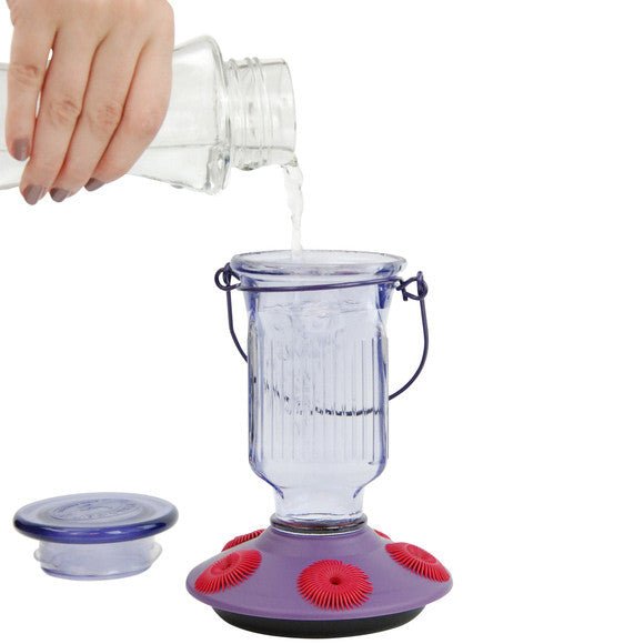 Hand pouring nectar into the Perky-Pet Lavender Field Top-Fill Glass Hummingbird Feeder 16 oz, featuring a wide-mouth opening and removable lid.